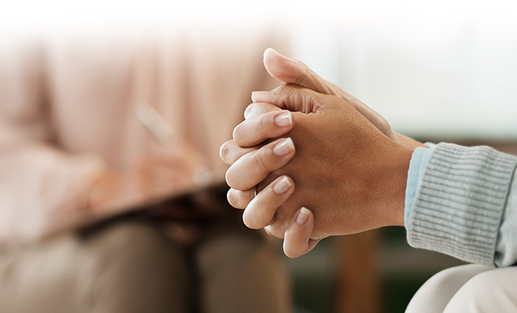 Patient's hands folded, therapist writing on clipboard in the background